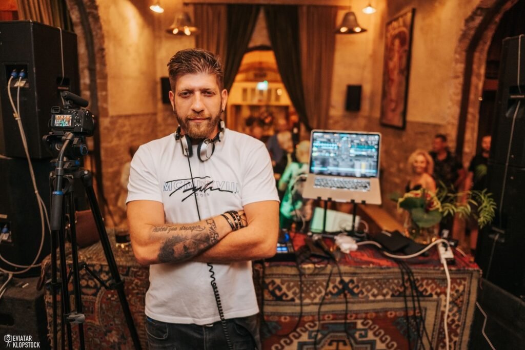 די.ג'יי עומד בעמדת דיג'יי באירוע ערב עם אוזניות סביב הצוואר וציוד סאונד מקצועי ברקע